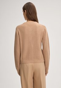 Donna con lunghi capelli castani che indossa un maglione di maglia beige e pantaloni beige, in piedi con le spalle rivolte verso l'osservatore su uno sfondo neutro.