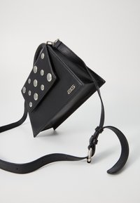 Sac à main en cuir noir avec un rabat texturé orné de clous ronds, une fine bandoulière ajustable, incliné pour une vue claire de son design.