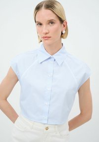 Camicetta azzurra chiara a maniche corte con bottoni, a righe verticali, colletto all'italiana e design senza cuciture per le spalle. Indossata con pantaloni bianchi a vita alta.