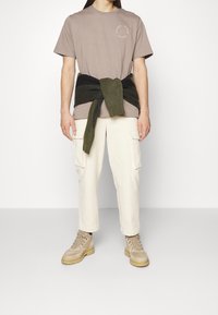 Světlé hnědé bavlněné tričko s malou grafikou, krémové cargo kalhoty a béžové šněrovací boty. Zelený svetr uvázaný kolem pasu.