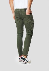 Olívovo zelené cargo rifle s viacerými vreckami, štíhlym strihom a textúrovaným povrchom. Nosené s bielymi a čiernymi športovými topánkami.