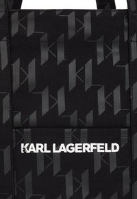 Sac fourre-tout en textile noir avec un motif gris "K" répété. Logo blanc "KARL LAGERFELD" sur le devant, avec des anses noires robustes.