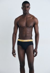 Svarta underkläder med en gul Nike-logga i midjan, med en tight passform och mjuk textur, bärs av en modell med atletisk fysik.
