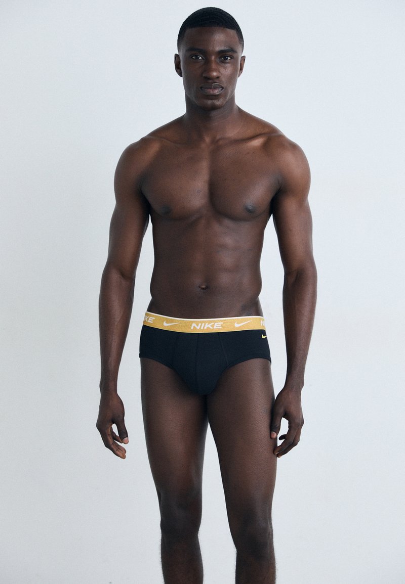 Svarta underkläder med en gul Nike-logga i midjan, med en tight passform och mjuk textur, bärs av en modell med atletisk fysik.