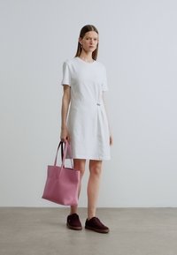 Λευκό φόρεμα με κοντά μανίκια και λεπτομέρεια συστροφής στο πλάι, συνδυασμένο με ροζ τσάντα χειρός και σκουρόχρωμα καφέ αθλητικά παπούτσια σε ανοιχτό γκρι πάτωμα.