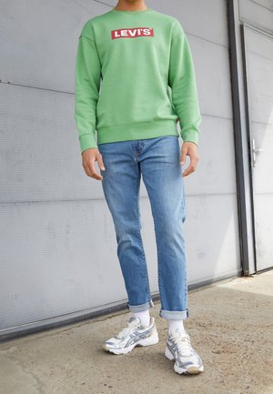 Person trägt grünes Levi's Sweatshirt, hochgekrempelte blaue Jeans, weiße Socken und silber-weiße Turnschuhe und steht auf Beton in der Nähe einer Metalltür.