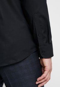 Czarna koszula z długim rękawem, z mankietem na guziki, o gładkiej fakturze i dopasowanym kroju. Noszona z granatowymi spodniami w kratę.