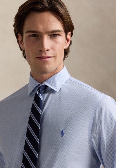 Lyseblå stripete skjorte med spiss krage, med en liten brodert logo på brystet; kombinert med et marineblått stripete slips.