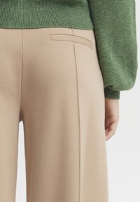 Béžové kalhoty s hladkým povrchem mají zadní kapsu a jemné švy, doplněné zeleným svetrém z ribbed pleteniny.