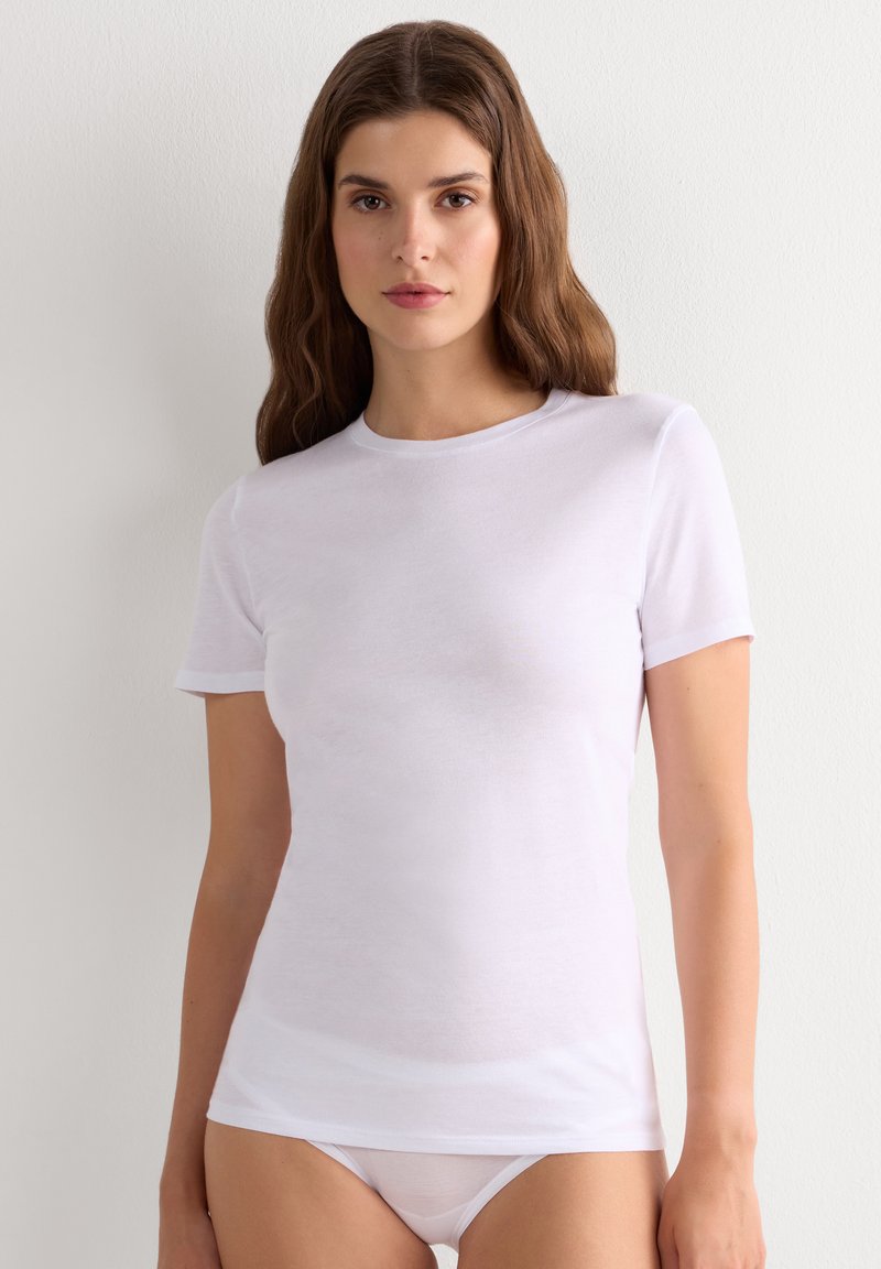 Kvinde med skulderlangt brunt hår iført en enkel hvid kortærmet t-shirt og matchende hvidt undertøj mod en neutral baggrund.