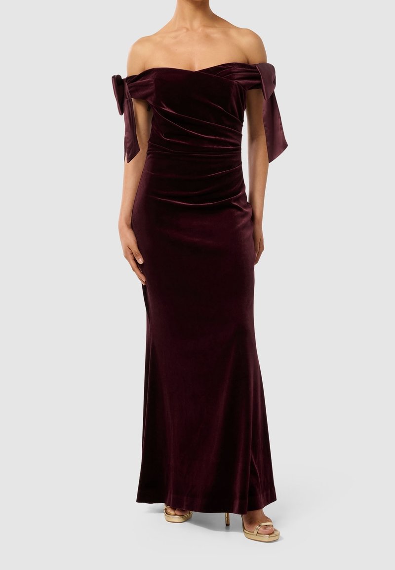 Femme vêtue d'une robe longue jusqu'au sol en velours violet foncé décolletée, avec des nœuds sur les manches et des talons ouverts dorés.