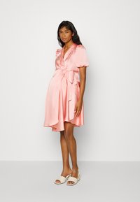 Vestido envolvente de cetim rosa com mangas bufantes, laço na cintura e bainha assimétrica. Usado com mules de salto bloco em bege claro. Textura suave.