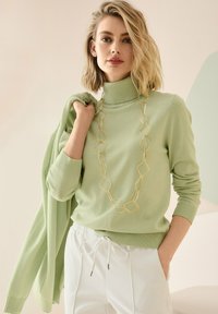 Blonde Frau mit einem hellgrünen Rollkragenpullover, die eine passende Strickjacke über der Schulter trägt, weiße Hose und eine lange goldene geometrische Halskette.