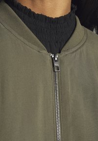 Olive green bomber jacket with a silver zipper, ribbed collar, and black ruffled inner lining. Smooth texture with a fitted design.