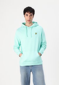 Mäta zelená mikina s kapucňou so šnúrkou a klokankou, s malým žltým vyšitým logom na ľavej hrudi.