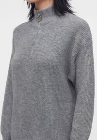 Person iført en grå strikket sweater med høj krave og halv lynlås med en guldring som lynlås.