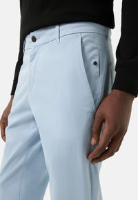 Lichtblauwe broek met een gladde stofstructuur, voorzakken en een knoopsluiting met een zwarte accent bij de zak.