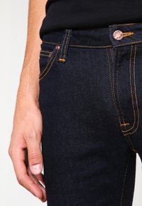 Mörkblå denimjeans med orange sömmar, med en framficka, knappstängning och slät textur. Närbild på midjebandet och hand.