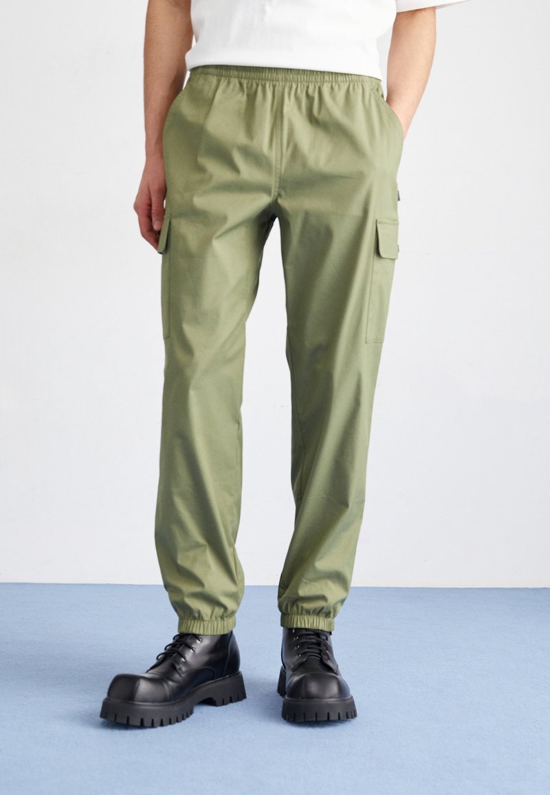 Personne portant un pantalon cargo vert olive avec des poignets élastiques et des bottes noires épaisses à lacets, debout devant un fond uni.