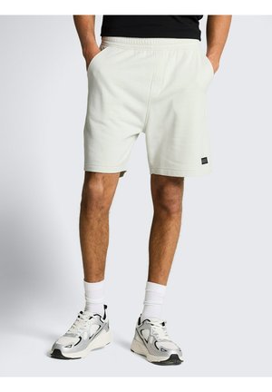 Man draagt witte sportshorts met zijzakken, witte kousen en witte en zwarte hardloopschoenen, staat met handen in de zakken.