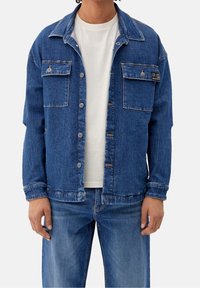 Chaqueta de mezclilla en azul medio, con mangas largas, dos bolsillos frontales, cierre de botones y un corte relajado. Camiseta blanca debajo.