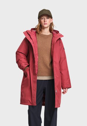 Personne portant un long manteau d'hiver rouge à capuche, un pull marron, un pantalon foncé et une casquette de baseball vert olive, debout devant un fond uni.