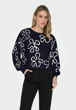 Marineblauer, verkürzter Pullover mit weißen Blumenmustern, runder Ausschnitt und langen Ärmeln. Texturiertes Strickmaterial. Kombiniert mit schwarzen Hosen.