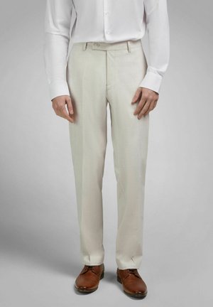 Mann trägt beige Anzughose, weißes Hemd mit Knöpfen und braune Lederschuhe, steht vor einem schlichten grauen Hintergrund.