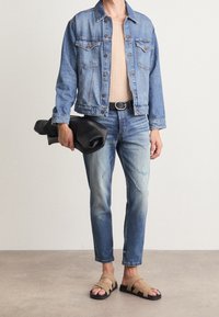 Chaqueta de mezclilla en azul claro, ajustada de punto beige debajo, jeans azul desgastados, cinturón negro, sosteniendo un bolso negro de mano y sandalias de tira color caqui.