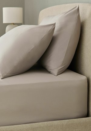 Zwei beige Kissen lehnen an einem beigen gepolsterten Kopfteil an einem Bett mit Spannbettlaken, mit einer weißen Tischlampe im Hintergrund.
