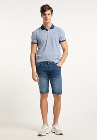 Polo azul de manga corta con patrón texturizado y ribete marino, combinado con pantalones cortos de mezclilla azul claro. El modelo lleva zapatillas blancas con acentos grises.