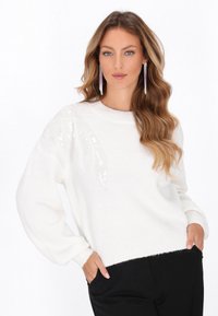 Pulover alb tricotat cu mâneci lungi, având un model texturat cu paiete pe un umăr. Material moale și detalii cu tiv ondulat.