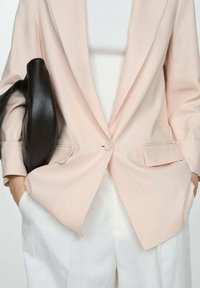 Light pink blazer made of soft fabric, single button closure, two front pockets, paired with a white top and loose white trousers, with a black handbag.