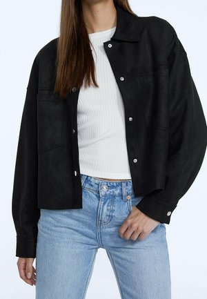 Femme portant une veste courte noire à boutons sur un haut blanc côtelé avec un jean taille haute bleu clair, devant un fond uni.