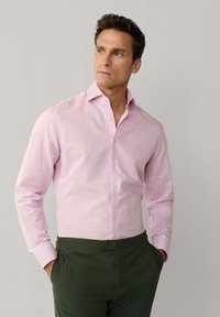 Mand iført en lyserød skjorte og mørkegrønne bukser, hænderne i lommerne, kigger let til siden mod en ensartet lys baggrund.