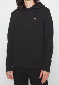 Sudadera con capucha negra con un patrón texturizado, que presenta un pequeño logo rojo y blanco en el pecho, y puños y dobladillo acanalados.