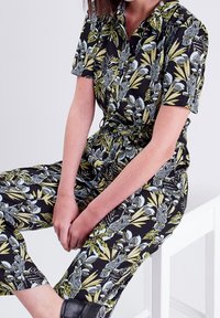 Combinaison à manches courtes en tissu noir, arborant un imprimé botanique vif en vert et bleu avec des oiseaux, et une taille ceinturée pour un ajustement ajustable.