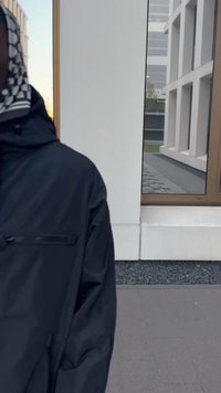 Sort hættetrøje med en glat tekstur, der har en lynlåslomme og et ternet tørklæde. Baggrunden inkluderer en moderne bygning og et reflekterende vindue.
