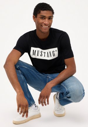 Sort bomulds T-shirt med et stort hvidt "MUSTANG" logo, parret med lyseblå denim jeans og hvide sneakers.