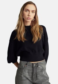 Černý zkrácený svetr s vysokým výstřihem a volnými rukávy, kombinovaný s šedými džíny s vysokým pasem, které mají lehké odřeniny a kovové detaily.