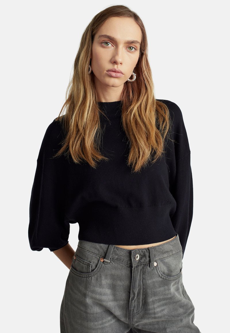 Černý zkrácený svetr s vysokým výstřihem a volnými rukávy, kombinovaný s šedými džíny s vysokým pasem, které mají lehké odřeniny a kovové detaily.