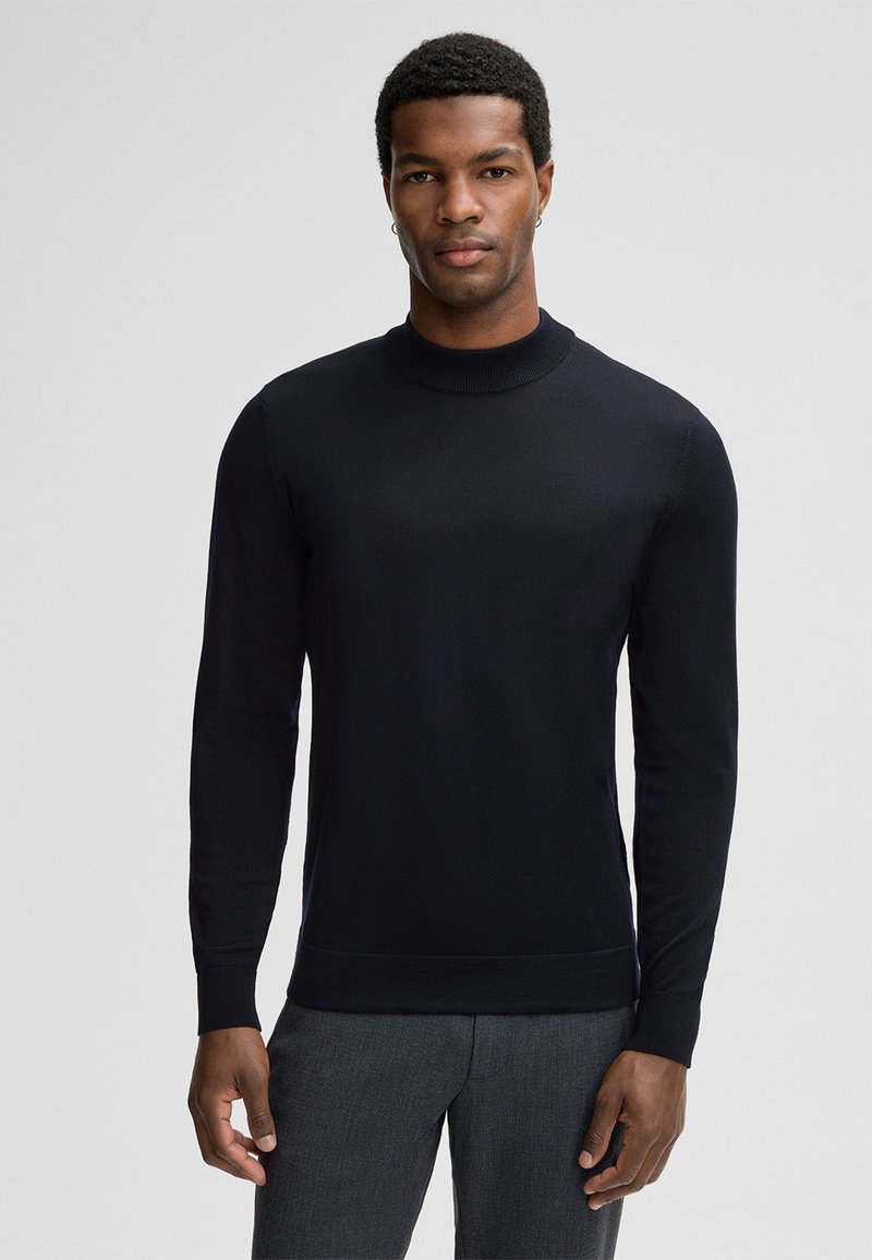 Marineblaue Rundhals-Strickjacke aus glattem Material. Eng anliegende lange Ärmel mit gerippten Bündchen und Saum. Getragen über grauen Hosen.