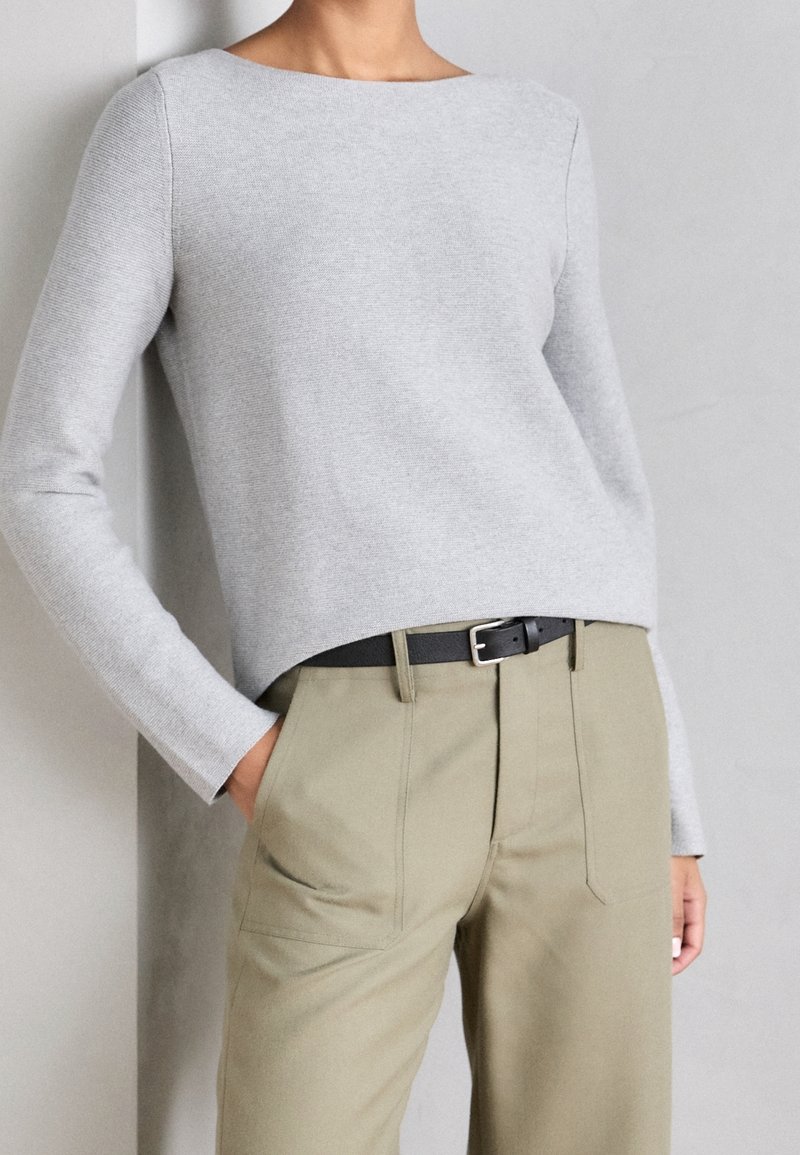 Henkilö, jolla on päällään vaaleanharmaa pitkähihainen paita, joka on työnnetty ylös korkeavyötäisiin beigeihin housuihin mustalla vyöllä, yksi käsi taskussa.