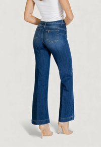 Jeans a zampa a vita alta di colore blu con tasche posteriori, realizzati in denim, caratterizzati da orli cuciti e una texture liscia. Indossati con tacchi alti beige.