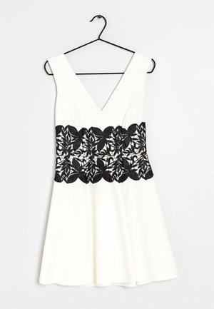 Witte mouwloze jurk met diepe V-hals, zwarte bloemenkantband rond de taille, hangend aan een zwarte hanger tegen een witte achtergrond.