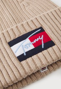 Bonnet en tricot beige avec une texture côtelée, arborant une vignette de logo multicolore avec des éléments bleus, rouges, blancs et noirs. Étiquette en bas visible.
