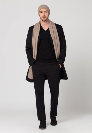 Manteau long noir, pull-over noir en v, pantalon noir, écharpe beige et bonnet assorti. Tous les articles ont des textures lisses et des coupes ajustées.