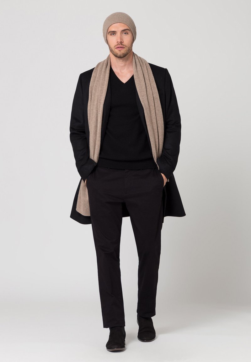 Cappotto lungo nero, maglione a collo a V nero, pantaloni neri, sciarpa beige e berretto coordinato. Tutti gli articoli hanno texture lisce e design aderenti.