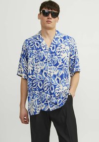 Camisa de manga corta azul y blanca con estampado floral, fabricada en un material ligero, con cierre de botones y diseño de cuello abierto. Combinada con pantalones negros.