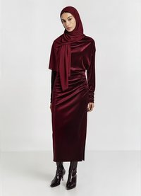 Burgunderfarbenes Satin-Kleid mit langen Ärmeln und seitlichem Schlitz, kombiniert mit einem passenden Hijab und glänzenden schwarzen kniehohen Stiefeln. Glatte Textur.